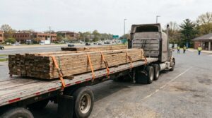 flatbed semi-truck hauling strapped lumber load on public roadway near workers